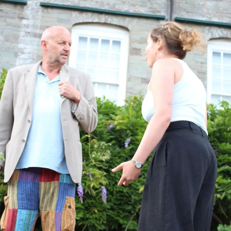 Two actors rehearsing an outdoor scene in front of a stone building.