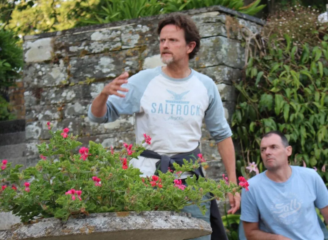 Two men rehearsing outdoors in a garden beside a stone wall and flowering urn