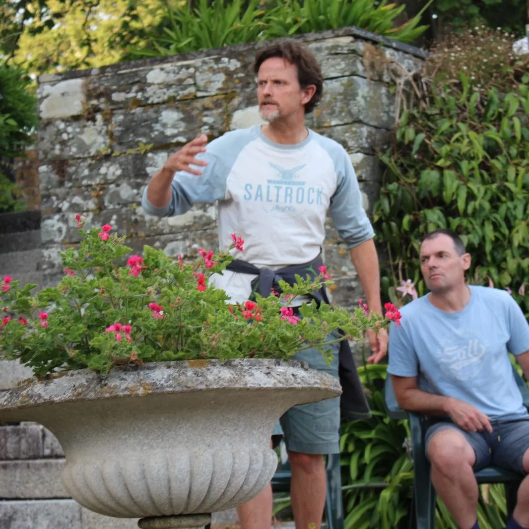 Two men rehearsing outdoors in a garden beside a stone wall and flowering urn