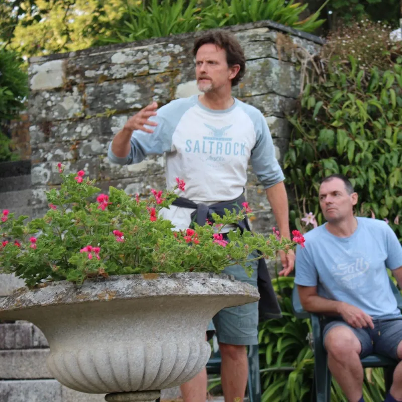 Two men rehearsing outdoors in a garden beside a stone wall and flowering urn