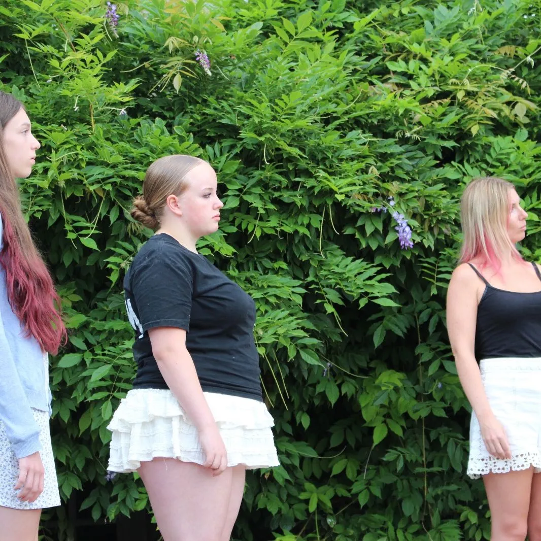 Three young women stand in profile during an outdoor rehearsal beside dense green foliage.