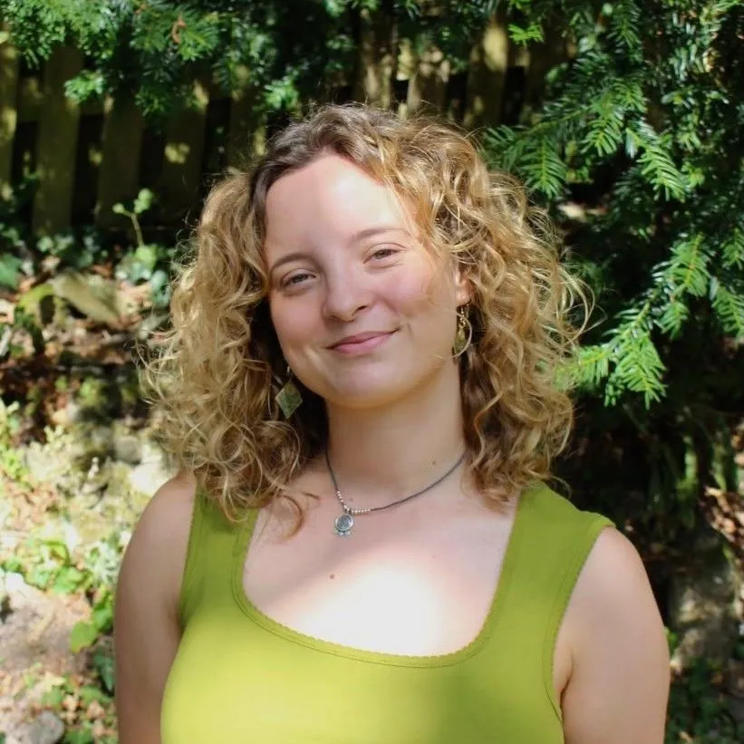 Portrait of a smiling woman with curly blonde hair standing beneath leafy trees