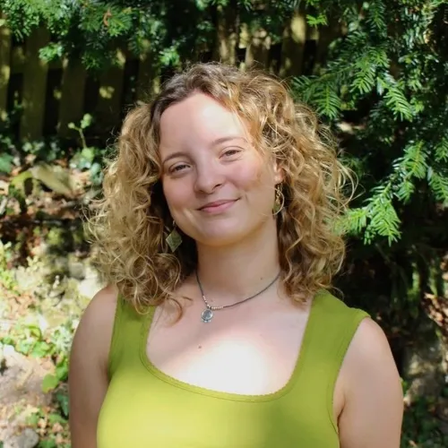 Portrait of a smiling woman with curly blonde hair standing beneath leafy trees