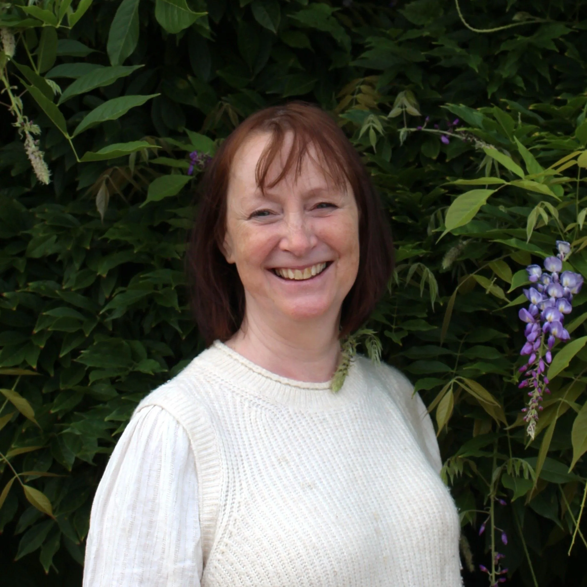 Smiling woman in a cream knitted vest standing beside purple flowers and leafy garden foliage.
