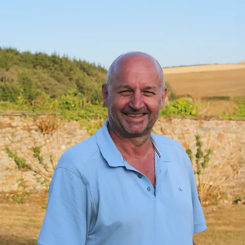 Smiling man in a light blue suit jacket with a blurred landscaped background.