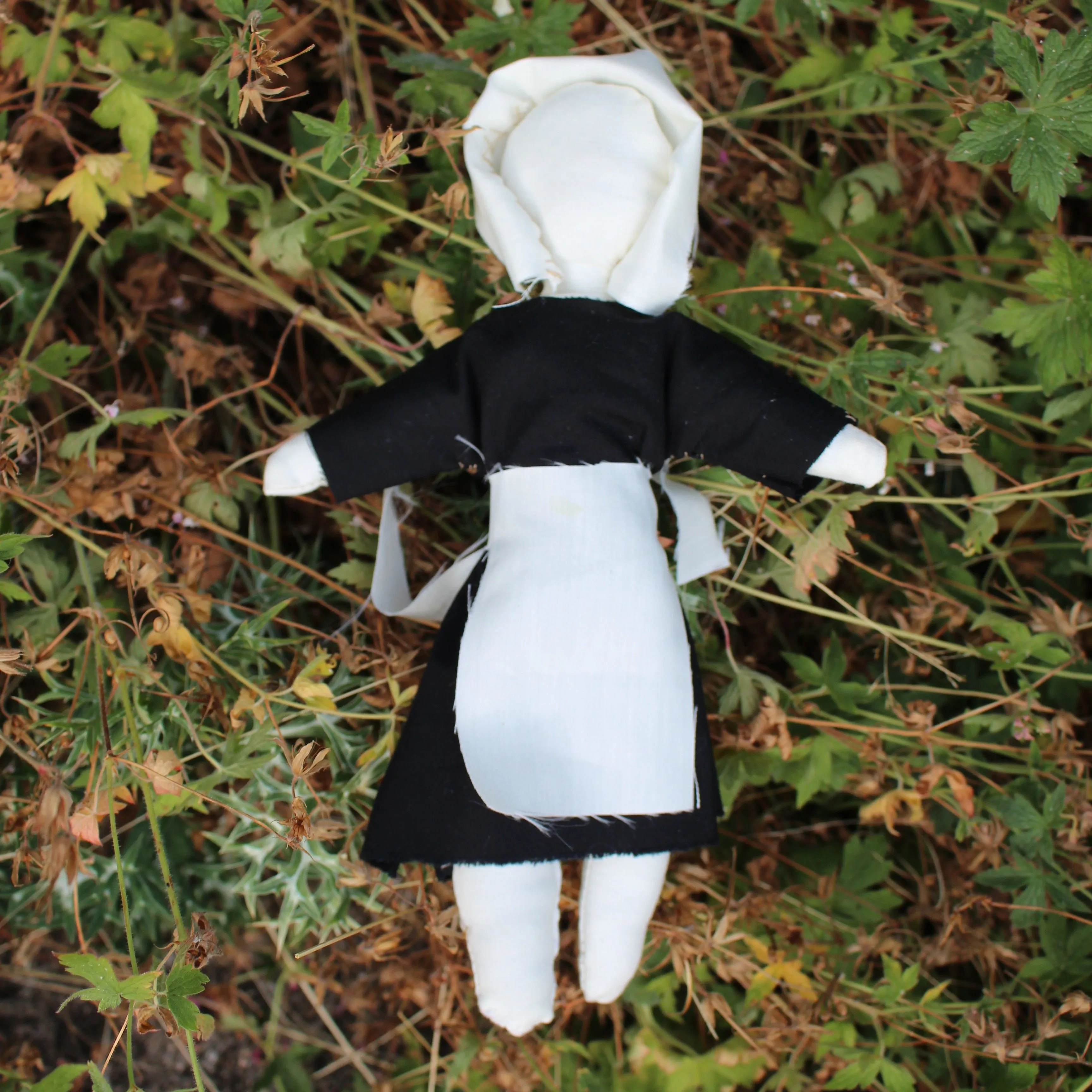 Handmade faceless cloth doll in a black dress with a white apron lying among wild plants