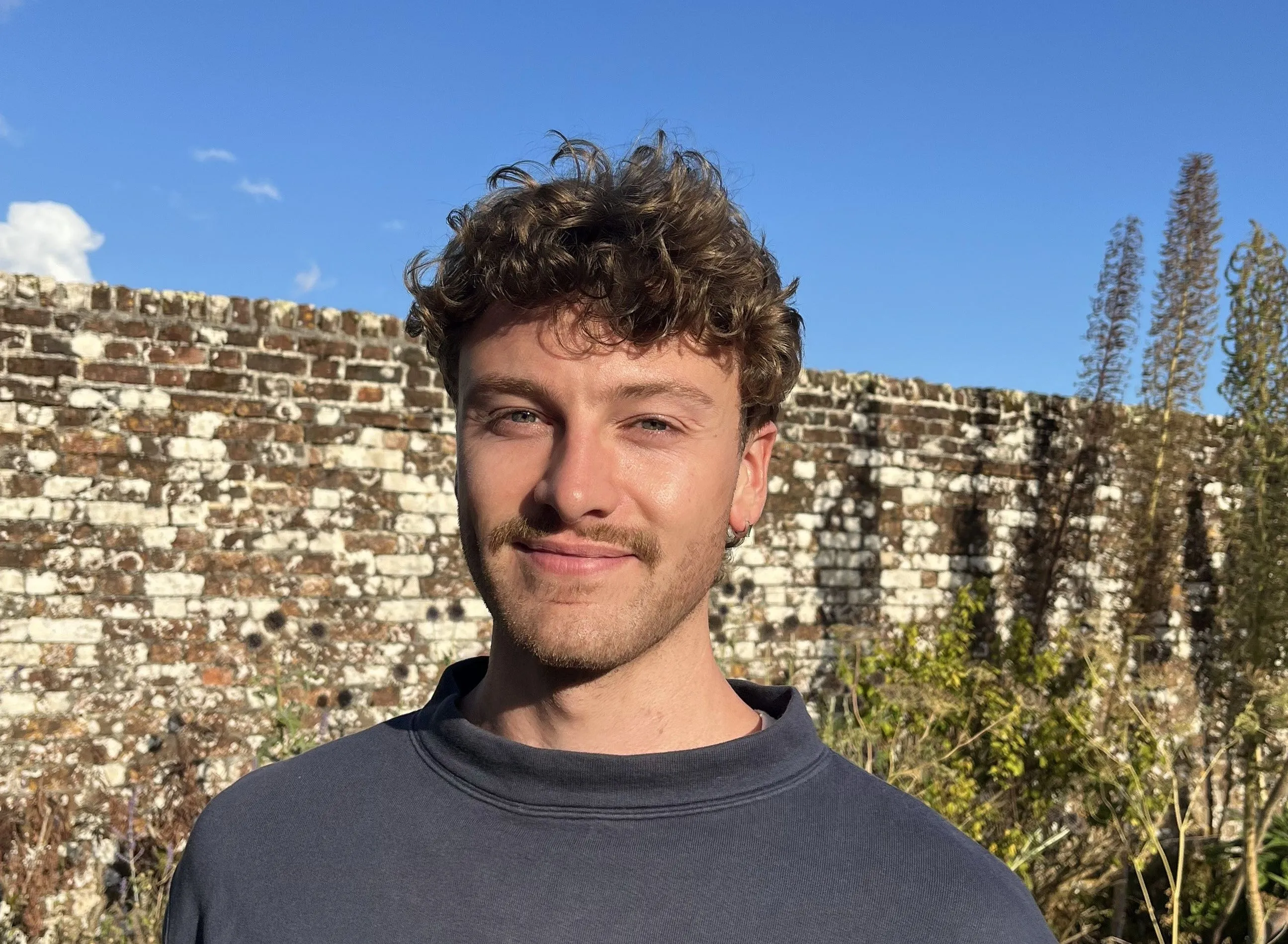 Portrait of a smiling young man with curly hair standing before an old brick wall under a clear blue sky.