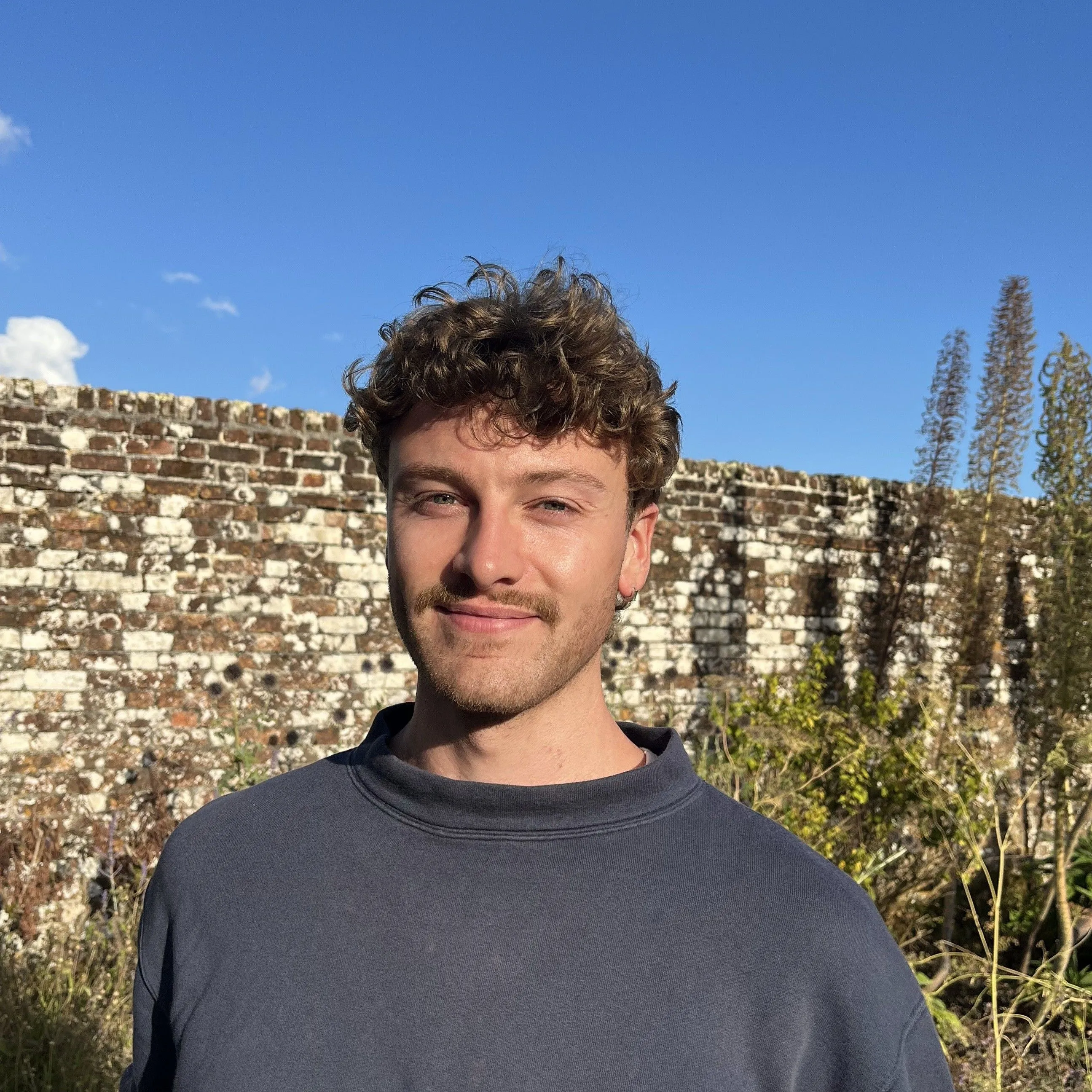 Portrait of a smiling young man with curly hair standing before an old brick wall under a clear blue sky.