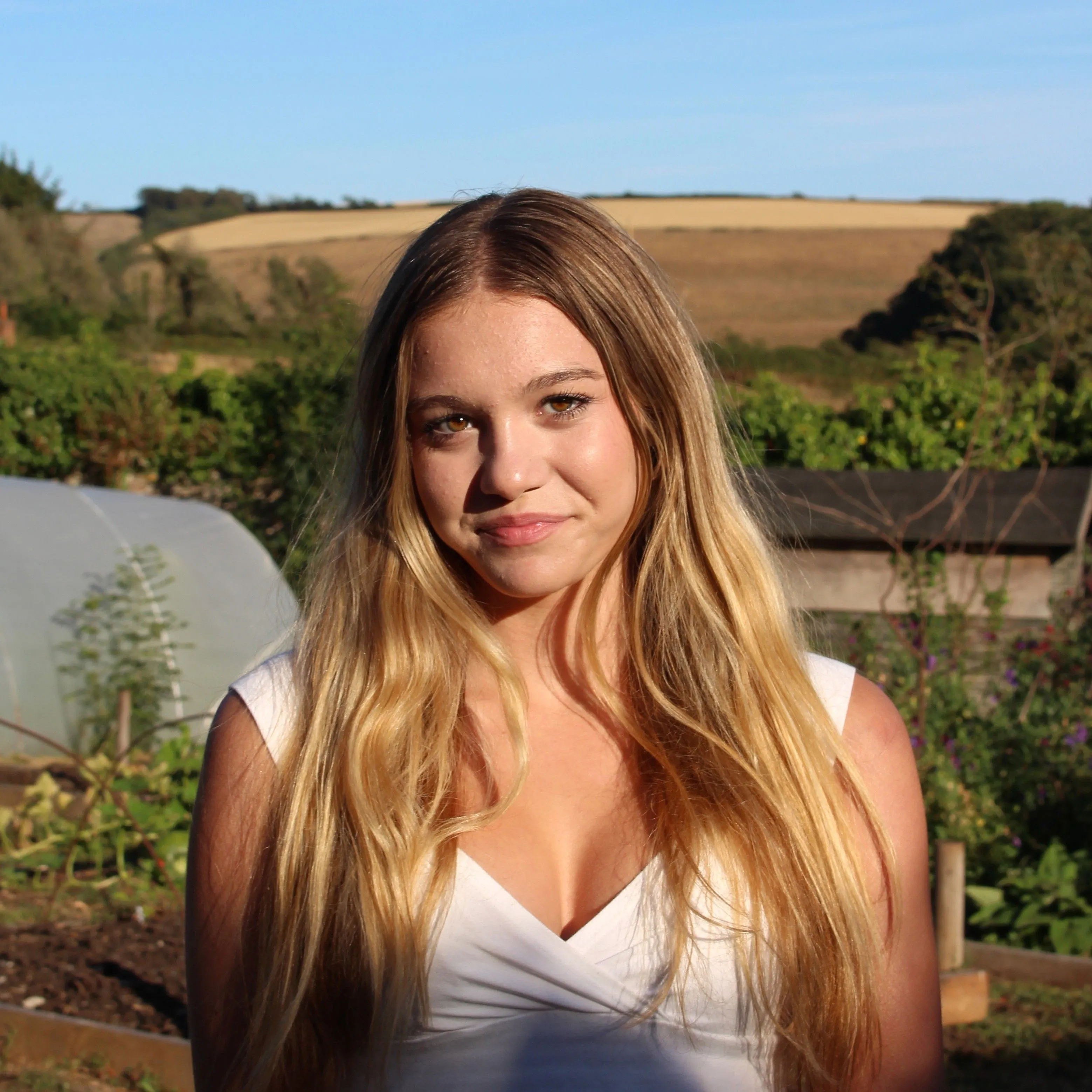 Sunlit portrait of a young woman with long blonde hair standing in a rural garden