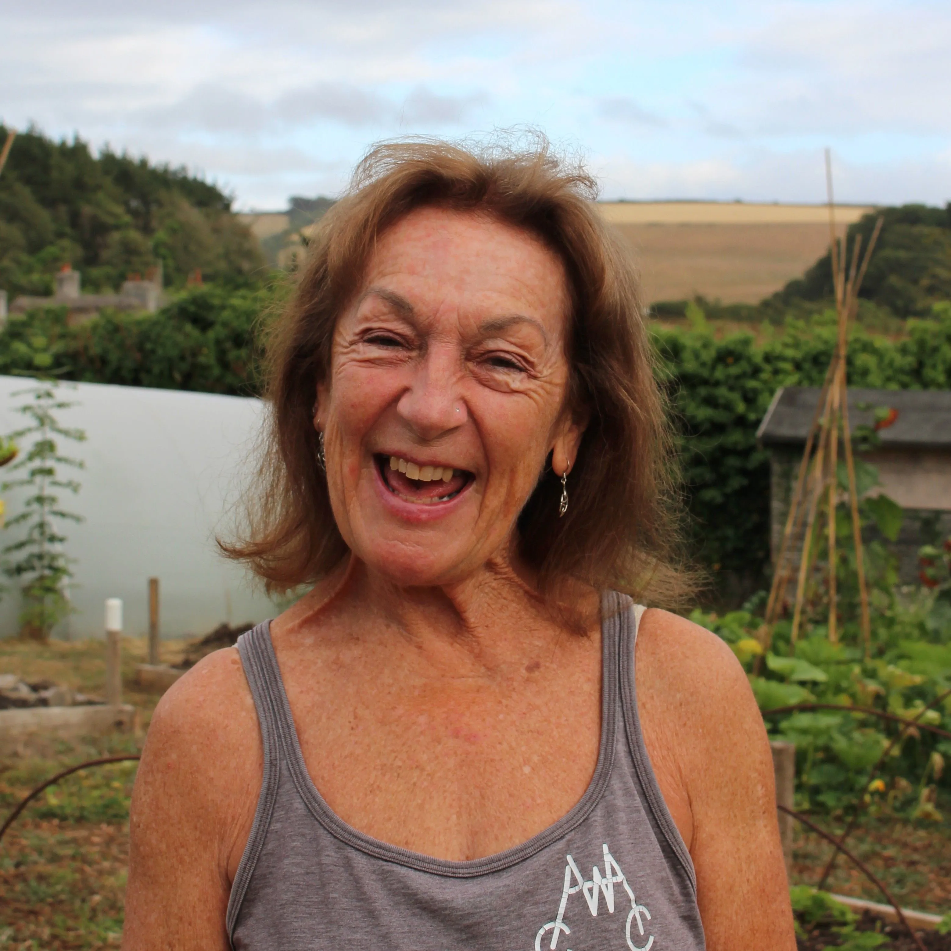 Smiling older woman standing in a vegetable garden with countryside fields behind her