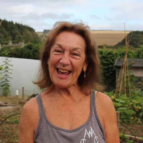 Smiling older woman standing in a vegetable garden with countryside fields behind her