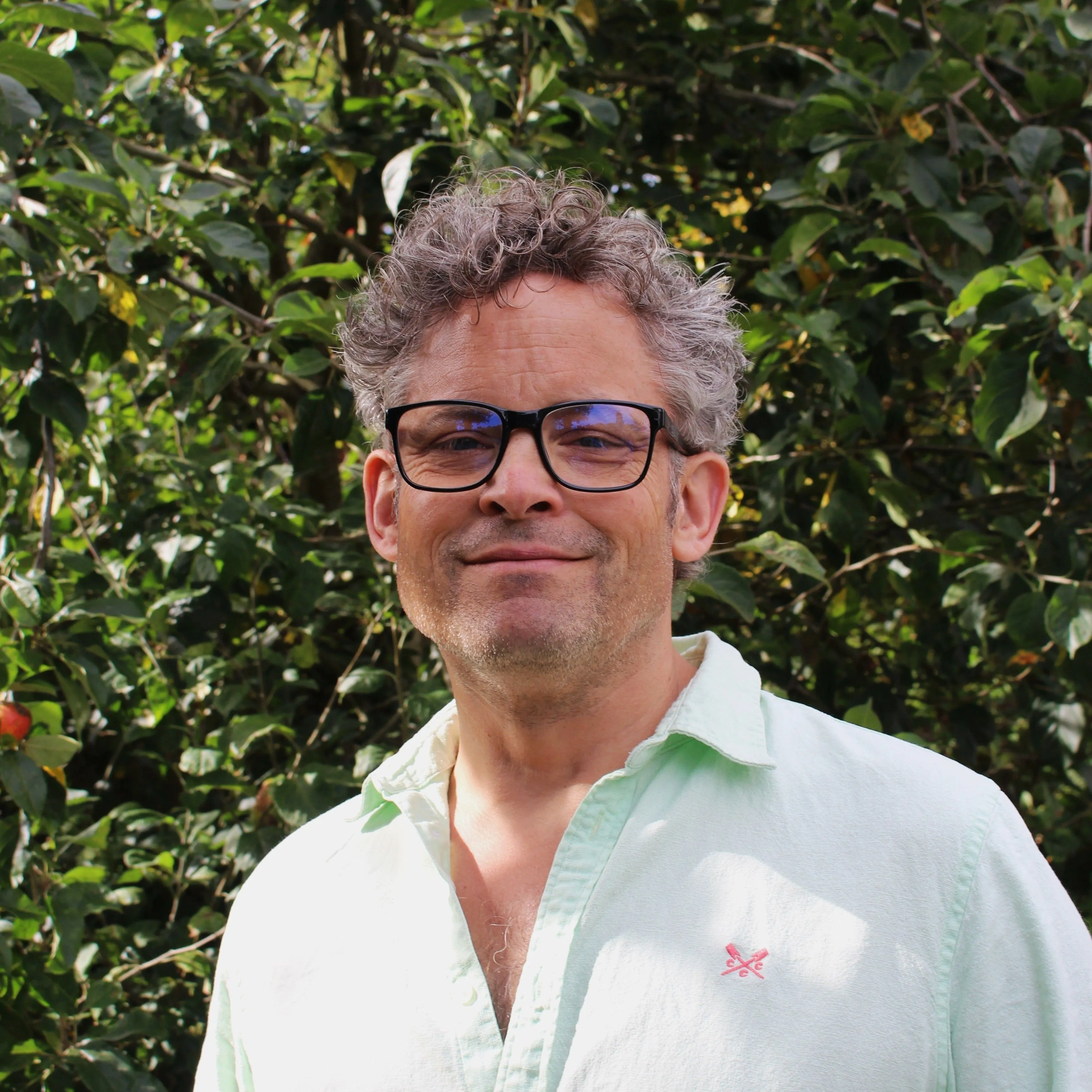 Portrait of Dave Fardon wearing glasses and a pale shirt in front of leafy greenery