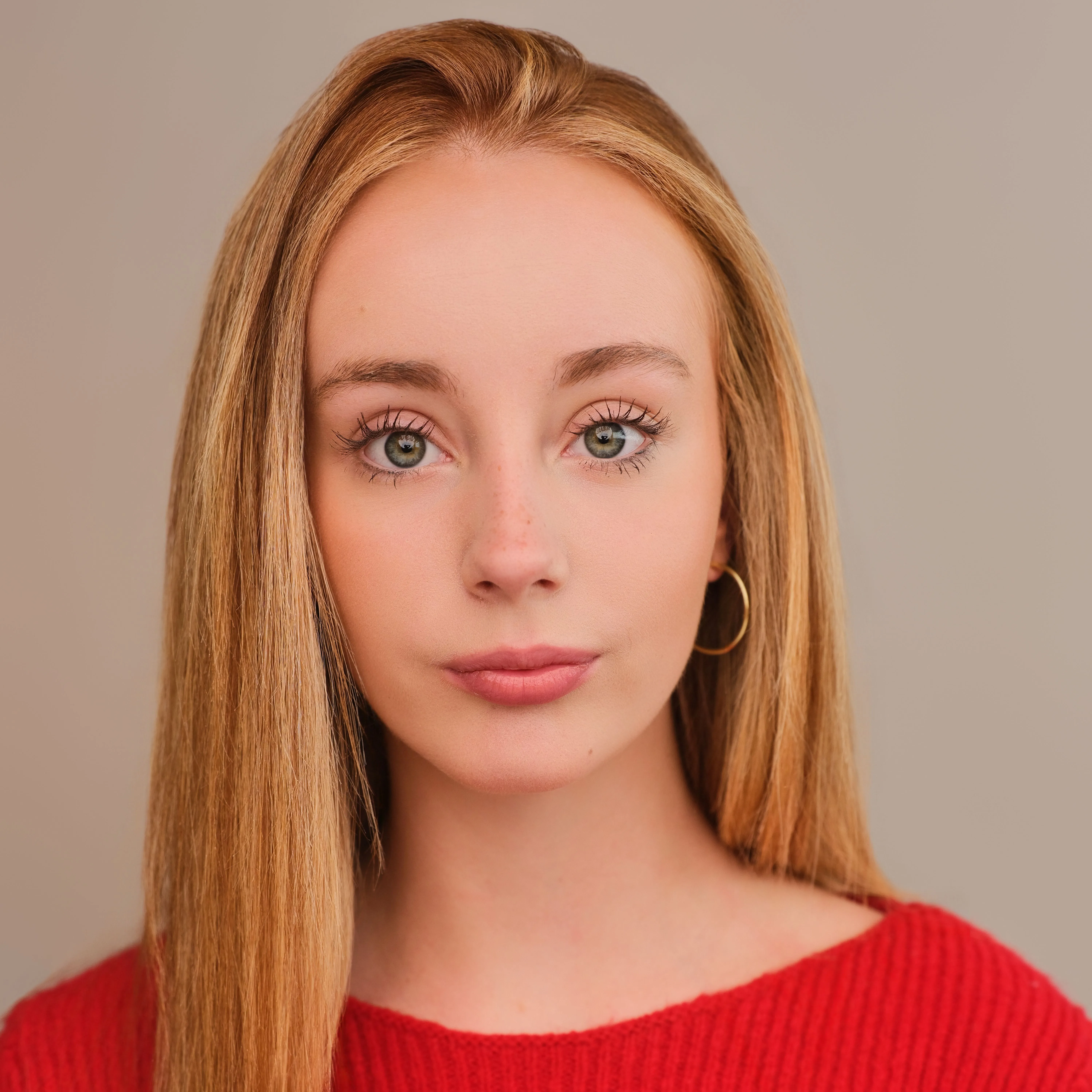 Close-up studio portrait of a woman with long blonde hair wearing a red jumper and gold hoop earring.