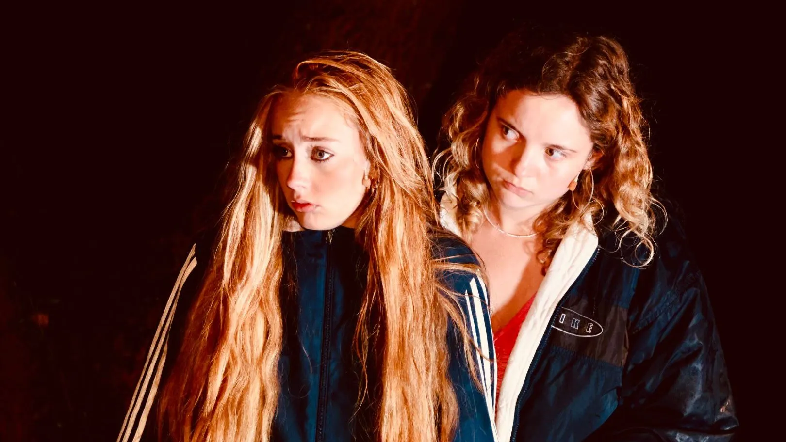 Two young women in dark sports jackets stand close together against a shadowy background.