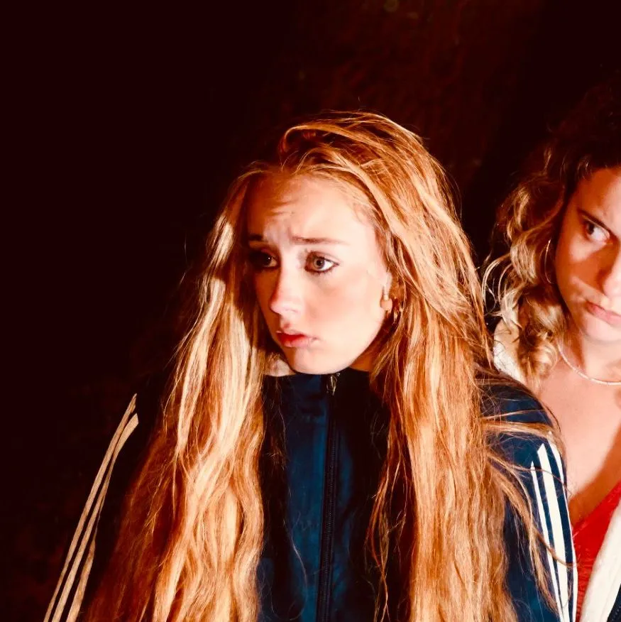 Two young women in dark sports jackets stand close together against a shadowy background.