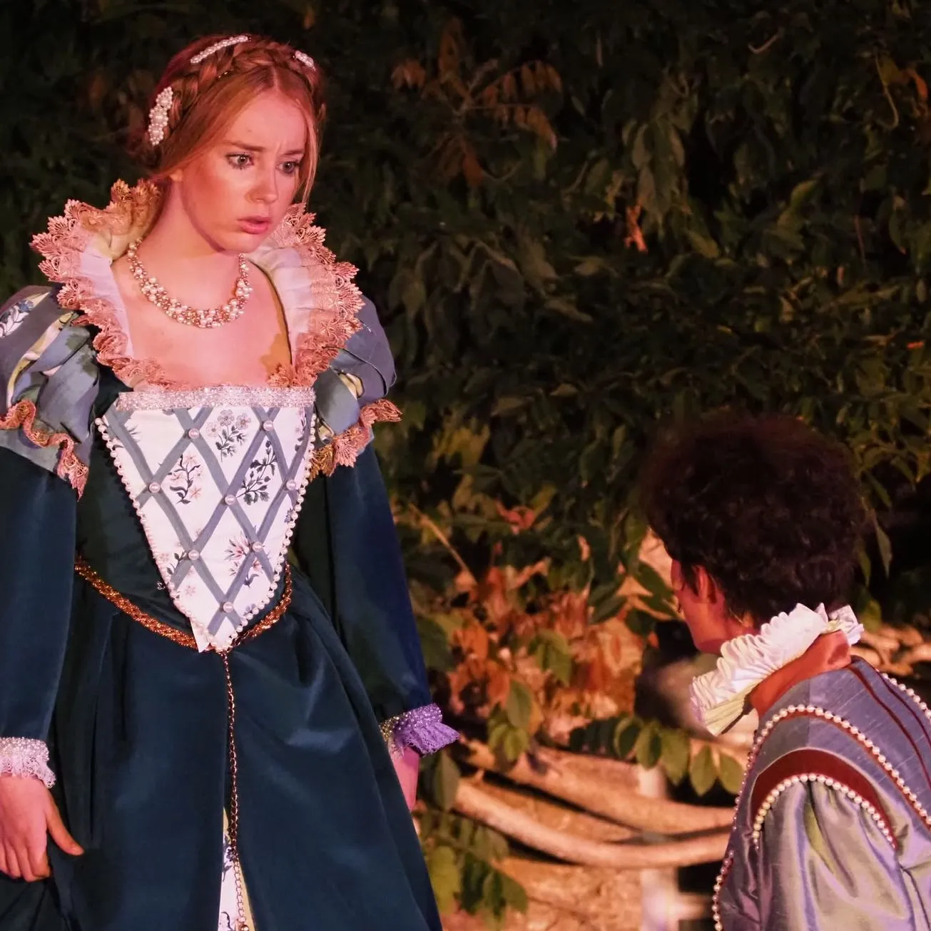 Actress in a dark green Renaissance-style gown performing outdoors opposite a kneeling costumed actor.
