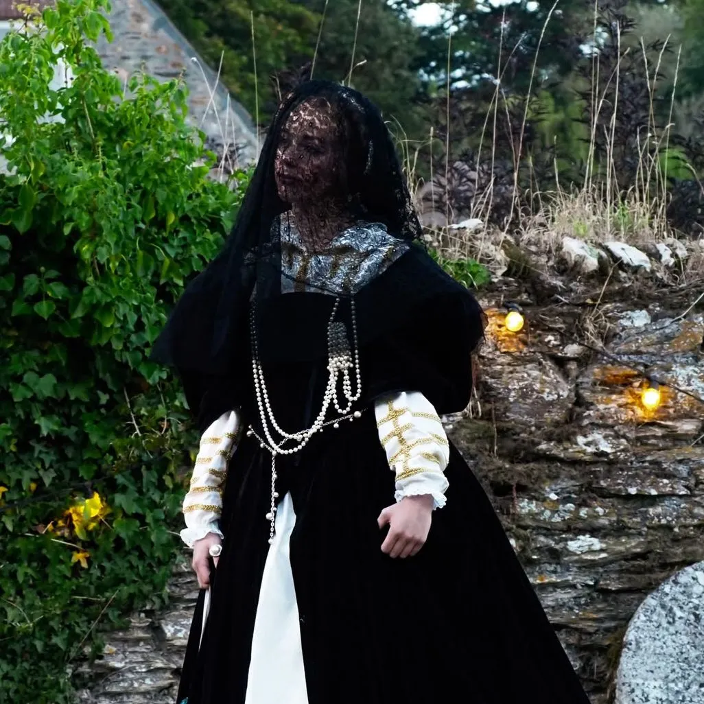 Two performers in historical costumes stand beside a stone wall and cottage garden, with one wearing a dark veil.