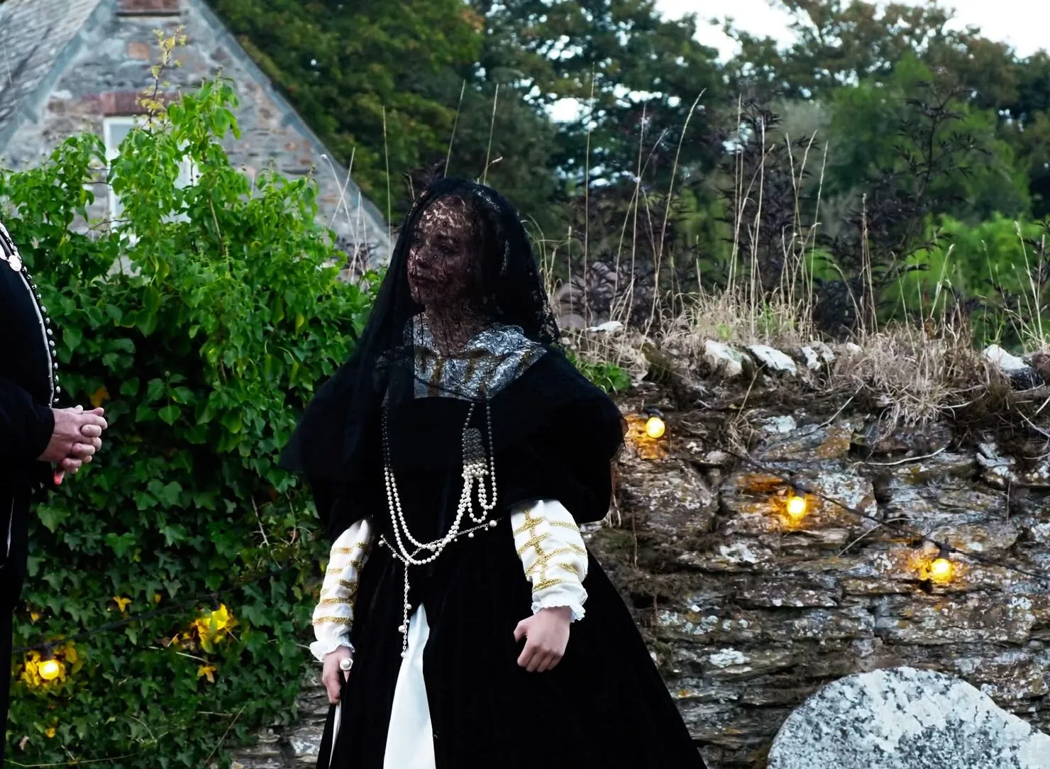 Two performers in historical costumes stand beside a stone wall and cottage garden, with one wearing a dark veil.