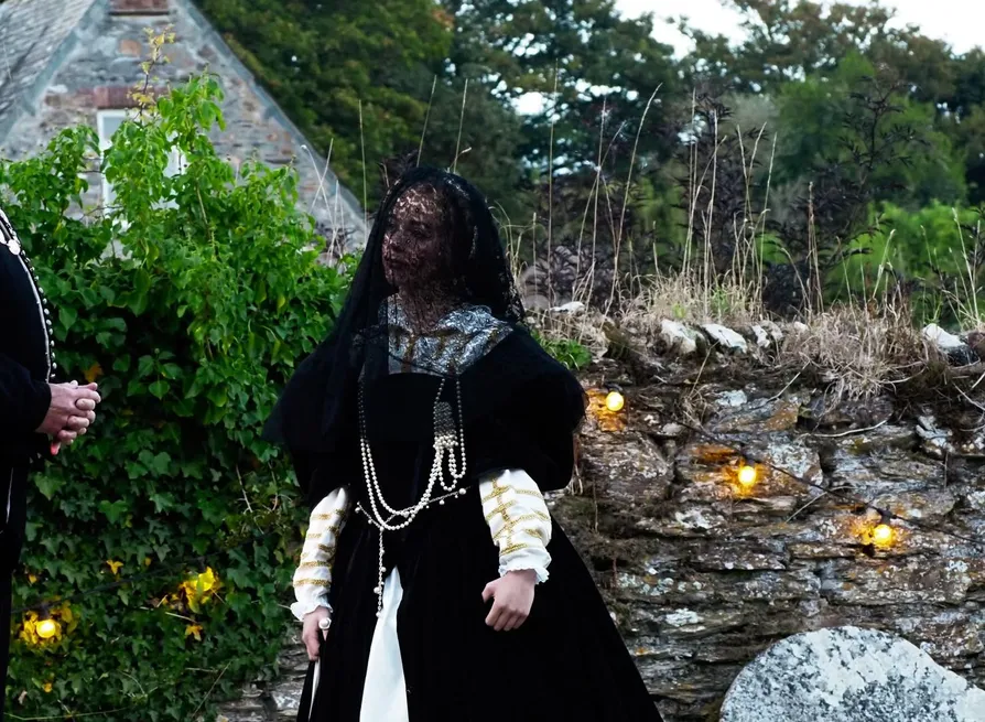 Two performers in historical costumes stand beside a stone wall and cottage garden, with one wearing a dark veil.