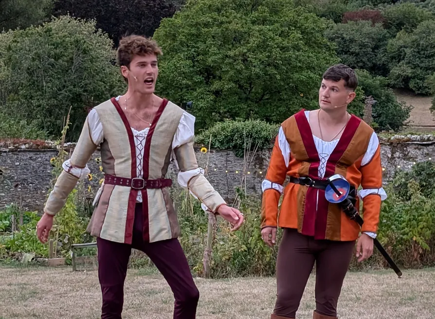 Two male actors in Renaissance-style costumes perform outdoors in a garden, one speaking while the other carries a sword.