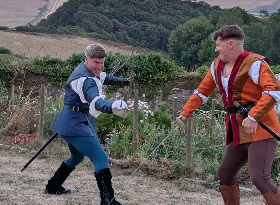 Two actors in colourful period costumes fencing outdoors against a backdrop of rolling countryside.