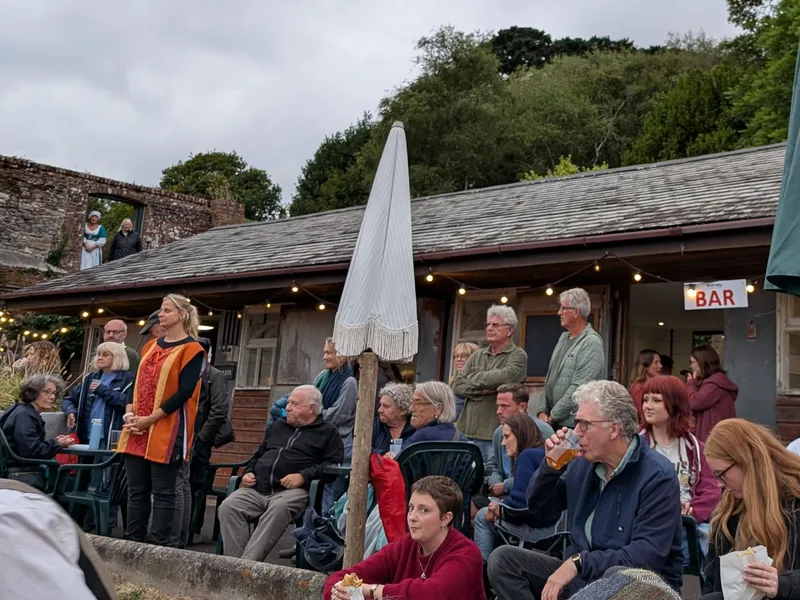 Audience gathered at an outdoor theatre venue beside a bar under an overcast sky.