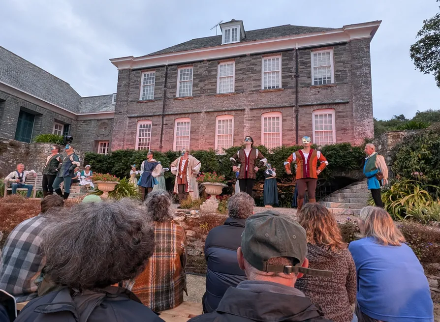 Audience watches a masked outdoor theatre performance in the garden of a historic stone house.