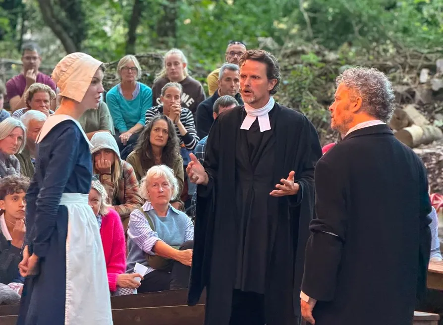 Outdoor theatre performance of The Crucible, with actors in Puritan costume before a seated woodland audience.