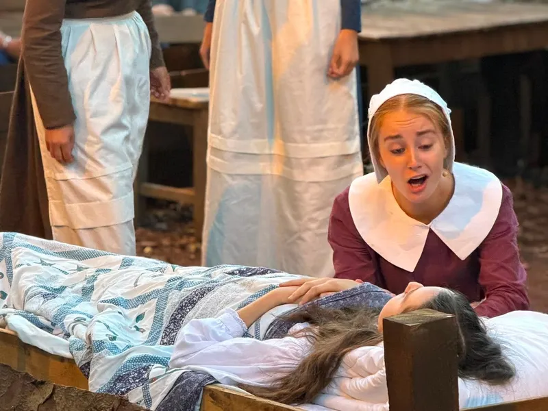 Actors in Puritan-style costumes perform a dramatic bedside scene on an outdoor stage.
