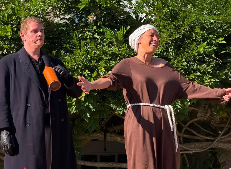 Two actors performing outdoors in period costume beside a stone building and garden foliage.