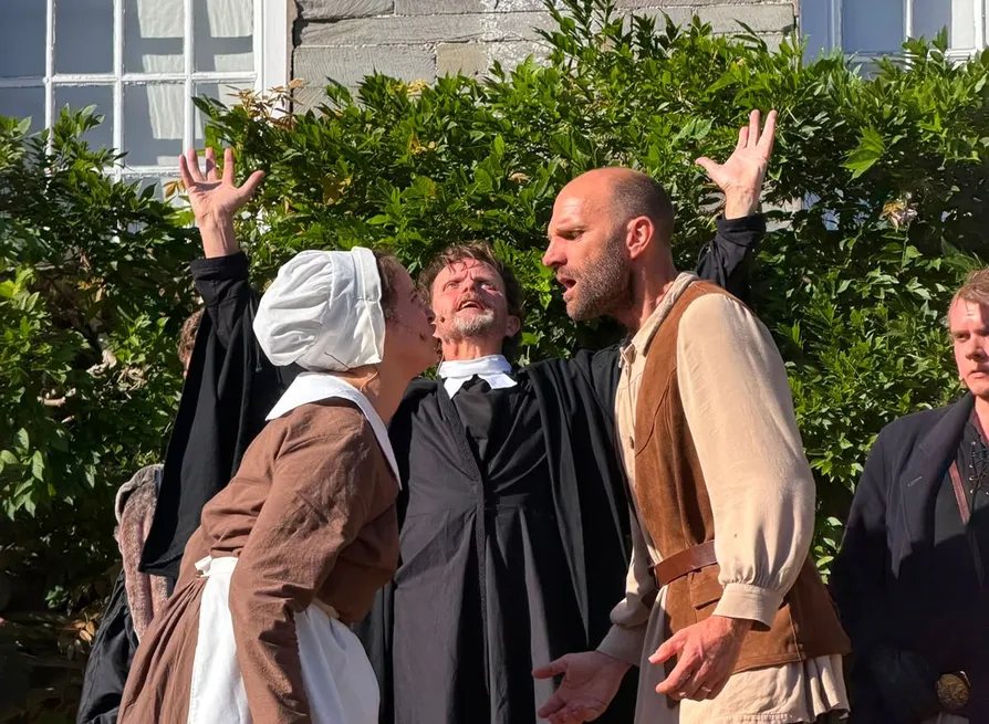 Outdoor theatrical performance with actors in period costumes, including a woman in a bonnet, a man in a waistcoat and a minister raising his arms.