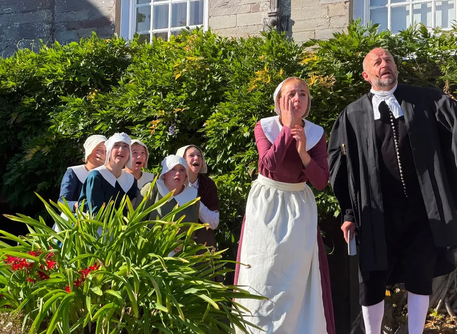 Actors in seventeenth-century costume react dramatically outside a stone house, with women in bonnets and a clergyman looking upwards.